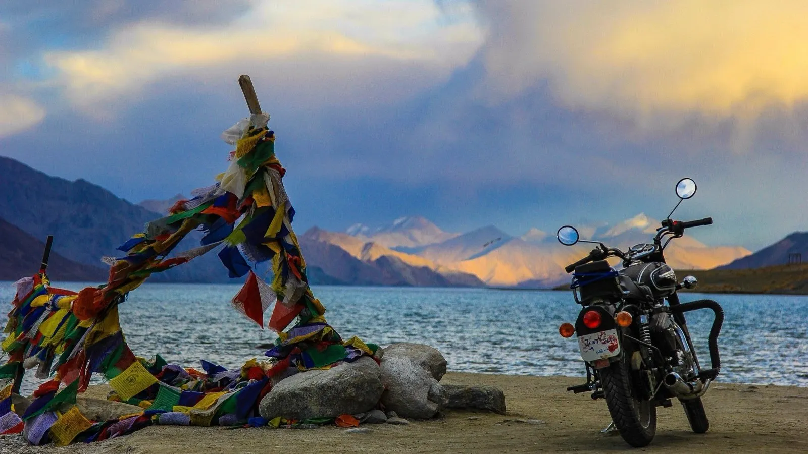 A Royal Enfield motorcycle parked beside strung prayer flags on the shore of Pangong Lake, with sunset light on the Ladakh peaks behind